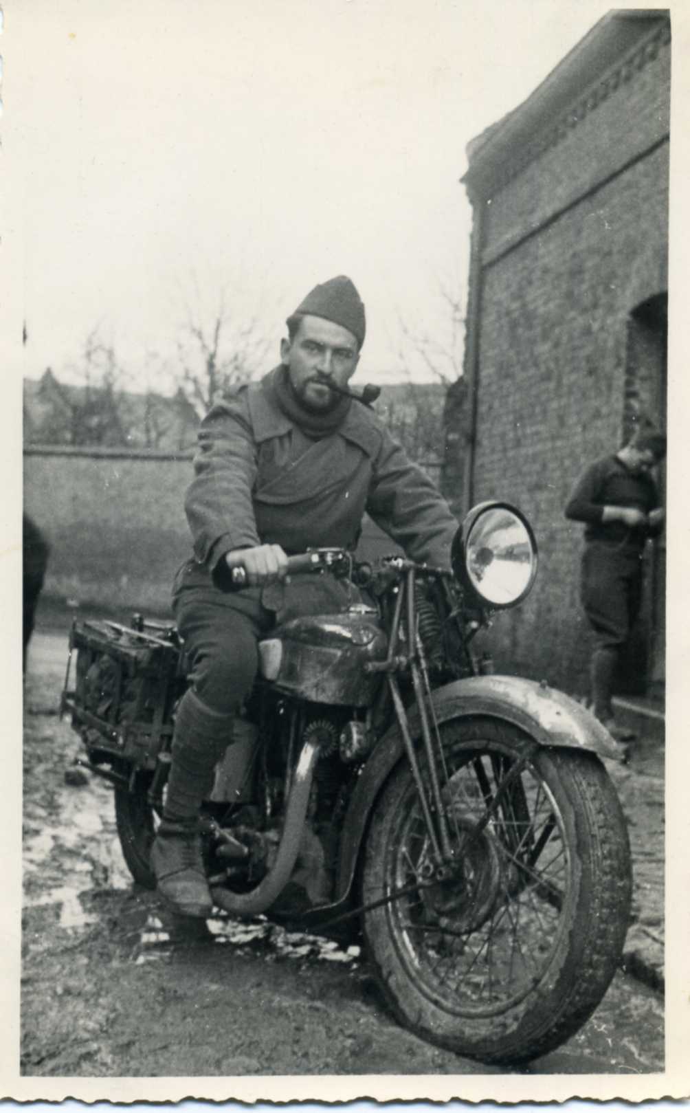 Pierre Roughol sits astride a motorcycle on a muddy road. He's a good-looking young man, 26 years old, brown hair and beard, and smokes a pipe. He wears a French military uniform, coat and cap.