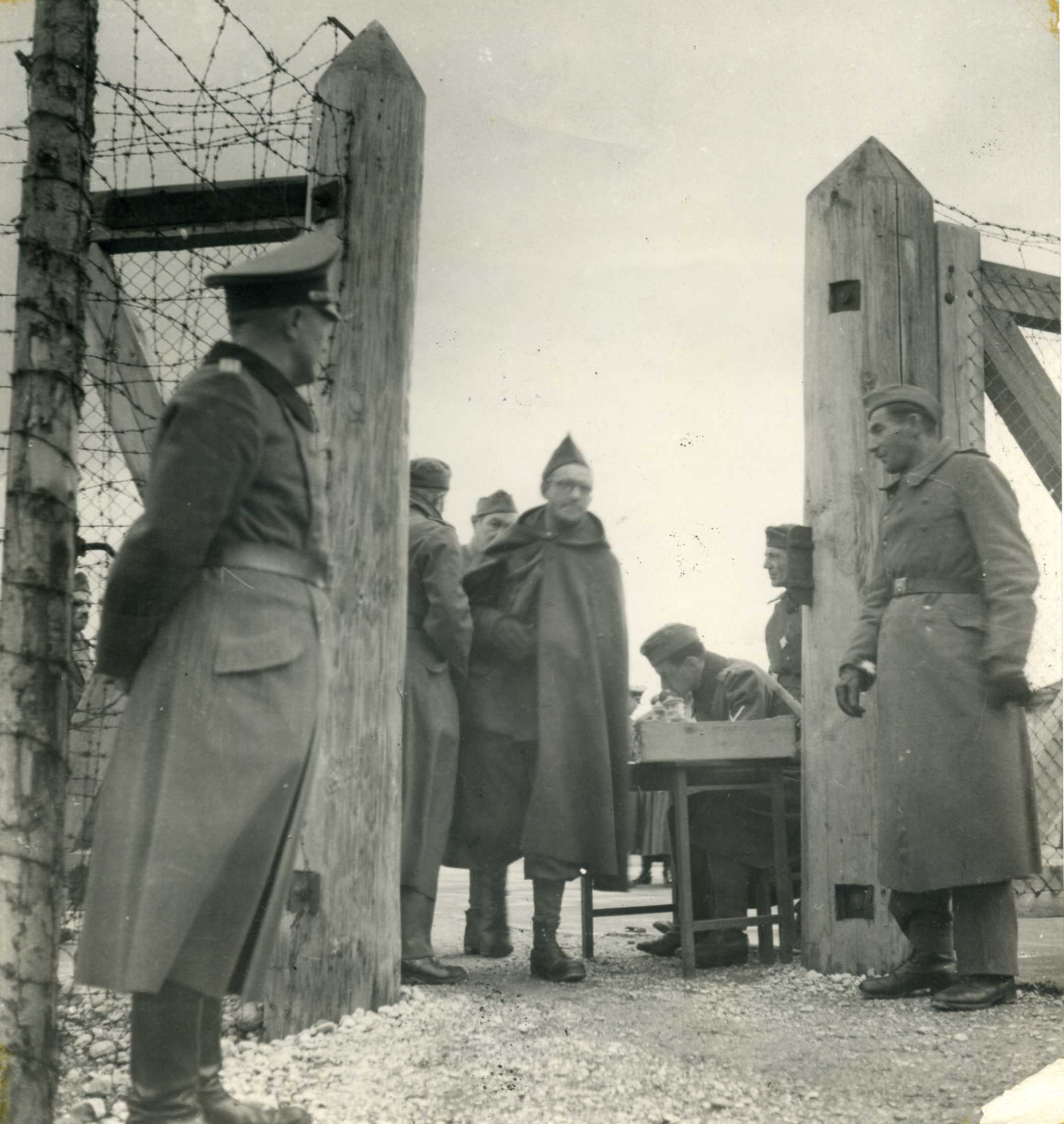 Black and white photograph. Gilbert Roughol, wearing a long cloak over a French military uniform, looks straight ahead as he steps passed a clerk at a desk and through the gate of the camp. He is framed by two tall wooden posts and fences of barbed wire. A German officer in uniform stands on the left, a French prisoner on the right, both looking at the man stepping away. 